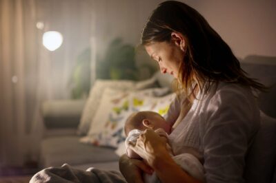 Young beautiful mother, breastfeeding her newborn baby boy at night, dim light. Mom breastfeeding infant