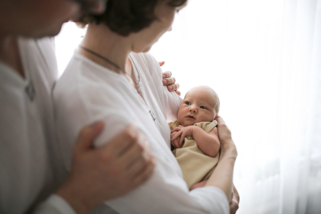 Husband-looking-over-wife's-shoulder-while-she-holds-newborn-baby
